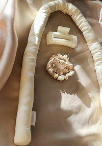 Kremowa, miękko wyściełana opaska na głowę, dopasowane gumki do włosów oraz duży klips do włosów ułożone na miękkiej beżowej tkaninie.