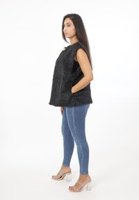 Czarna kamizelka z faux futra o luźnym kroju, z bocznymi kieszeniami i metalicznym akcentem, w połączeniu z niebieskimi obcisłymi dżinsami i przezroczystymi sandałami na obcasie.