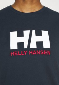 Camiseta de algodón azul marino con un gran logo blanco "HH" y el texto "HELLY HANSEN" en rojo, bordado en el centro. Diseño simple y casual.