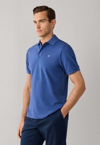 Homme debout les mains dans les poches, portant un polo bleu à manches courtes et un pantalon bleu foncé sur un fond uni.