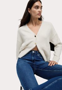 Krämfärgad stickad cardigan med V-hals, två framfickor och guldknappar, ihop med högmidjade blå jeans. Slät textur, avslappnad passform.