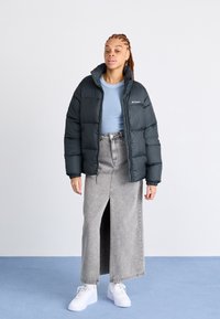 Giacca a vento nera con polsini elasticizzati, indossata sopra un top azzurro chiaro e una gonna di jeans grigia con uno spacco frontale. Sneakers bianche completano il look.