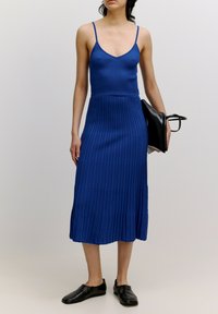 Blaues geripptes Strickkleid mit dünnen Trägern, das einen taillierten Oberteil und einen ausgestellten Rock hat. Begleitet von einer schwarzen Handtasche und flachen schwarzen Schuhen.
