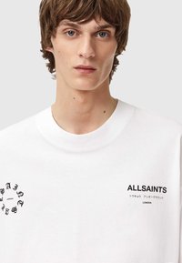 Valge puuvillane T-särk, millel on ümmargune kaelus, mustade graafiliste trükistega rinnal ja vasakul küljel. Sile tekstuur.