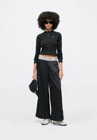 Μαύρο ριμπ μακρυμάνικο crop top με κουμπιά, μαύρο παντελόνι φαρδιάς γραμμής με λευκή ζώνη μέσης, γυαλιά ηλίου και μαύρο καπέλο. Αθλητικά παπούτσια με λεπτομέρειες.