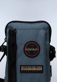 Petit sac bandoulière en tissu bleu avec fermeture éclair, présentant un patch logo circulaire et une étiquette de marque tissée avec des accents noirs.