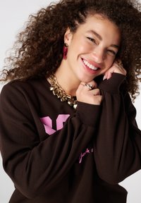 Bruine sweatstof met roze afbeelding, met lange mouwen. Gouden ketting en hartvormige oorbellen voegen accenten toe. Krullend haar zichtbaar.