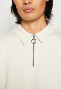 White zip-up pullover with a textured, patterned fabric and a flat collar. Features a silver zipper with a round pull tab.