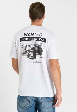 Hombre que lleva una camiseta blanca con la estampación en la espalda de la fotografía policial de un perro etiquetada como "WANTED MOST FUNNY DOG" y texto de advertencia debajo de la foto.