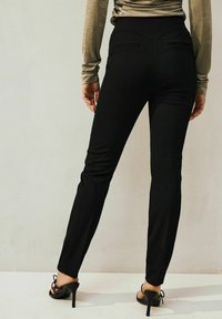 Černé kalhoty na míru s vysokým pasem, bočními kapsami a slim fit. Materiál vypadá hladce s jemným leskem. Nosené s černými sandály na podpatku.