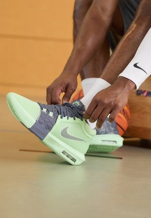 Persona atándose unas zapatillas de baloncesto Nike de color verde claro y gris, sentada en el suelo del gimnasio con una pelota de baloncesto naranja cerca.