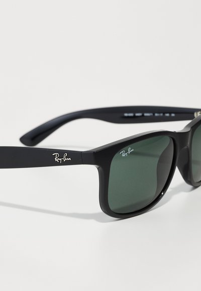 Lunettes de soleil en plastique noir avec des verres vert foncé. Présentent un logo sur la branche et un design rectangulaire avec une texture lisse.