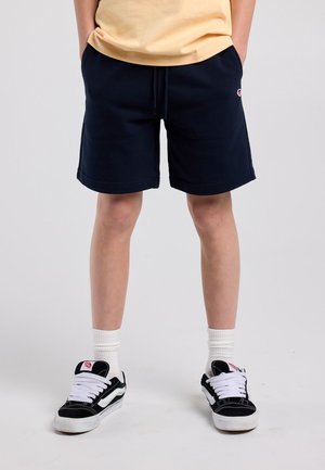 Giovane persona con pantaloncini neri, calzini bianchi, scarpe da ginnastica nere e bianche, in piedi con le mani in tasca davanti a uno sfondo semplice.