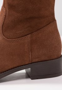 Brązowy zamszowy botek za kostkę na słupku, z kontrastowym przeszyciem i gładkim, zaokrąglonym kształtem. Teksturowana powierzchnia i delikatne załamania.