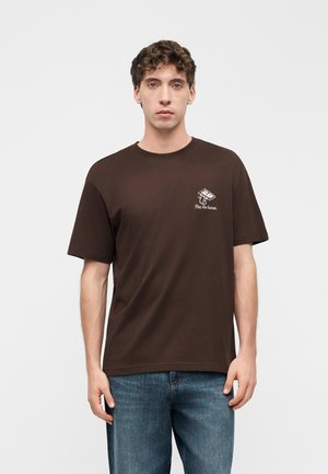 Jeune homme portant un t-shirt marron avec un motif de cassette et le texte « Play the future », associé à un jean bleu, debout sur un fond blanc.