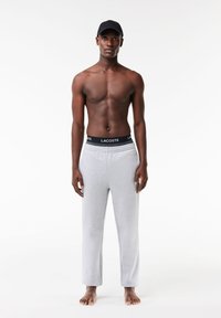 Lichtgrijze sweatpants met een zwarte Lacoste-logo tailleband. Gladde textuur, rechte snit, zonder zakken, gedragen op blote voeten, met een zwarte pet.