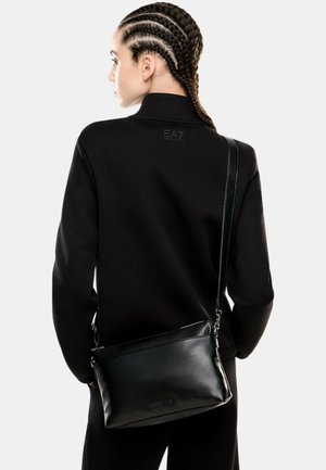 Borsa a tracolla in pelle nera con texture liscia, chiusura superiore con zip e tracolla regolabile, accompagnata da una felpa nera semplice.