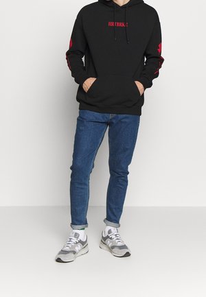 Persoon staand, gekleed in een zwarte hoodie met rode tekst en patches, blauwe opgerolde spijkerbroek, witte sokken en grijze sneakers.