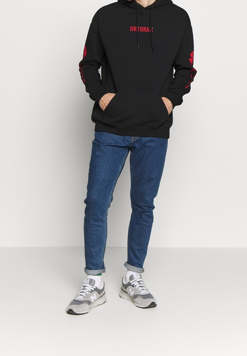 Personne debout portant un sweat à capuche noir avec texte rouge et écussons, jean bleu retroussé, chaussettes blanches et baskets grises.