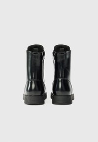 Paire de bottines noires brillantes à fermeture éclair latérale, semelles de marque et doublure intérieure rembourrée, vues de dos sur un fond uni.