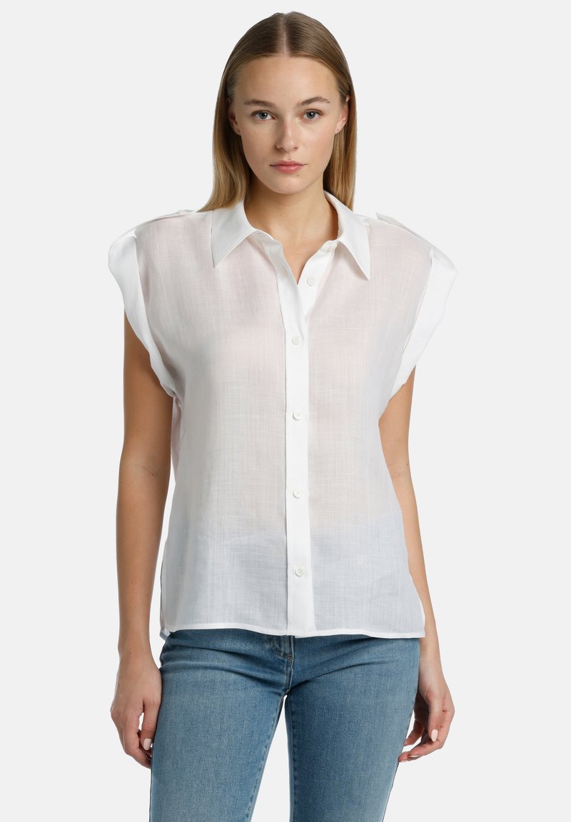 Blusa blanca sin mangas con frente de botones, que presenta un collar, tela transparente y una textura sutil; combinada con jeans de color azul claro.