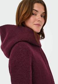 Manteau en laine bordeaux avec une grande capuche structurée, présentant une surface texturée et des poches sur les coutures latérales.