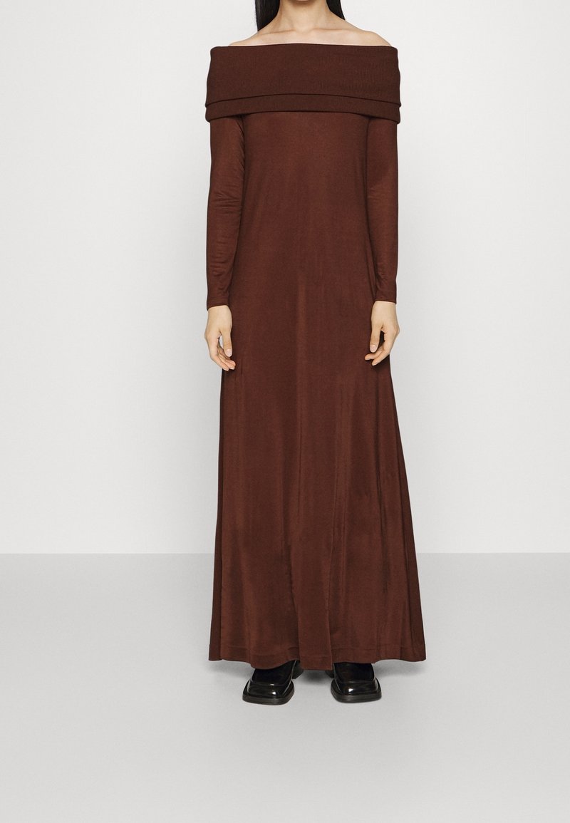 Lång brun off-shoulder klänning i ett mjukt, stretchigt tyg, med långa ärmar och en flödande silhuett, med svarta skor synliga nedanför.