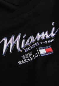 Μαύρο ύφασμα με λαμπερό κείμενο «Miami 2026 1-3 Μαΐου», λογότυπο της ομάδας Formula 1 της Cadillac και λογότυπο σημαίας Tommy Hilfiger τυπωμένα από κάτω.