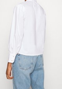 Personne portant un blouse blanche à manches longues avec des poignets en scallop et un jean taille haute bleu clair, de dos contre un fond uni.