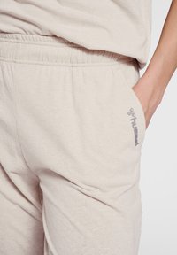 Light beige cotton blend joggers with an elastic waistband, side pocket, and subtle logo print in gray. Soft texture with slight sheen.