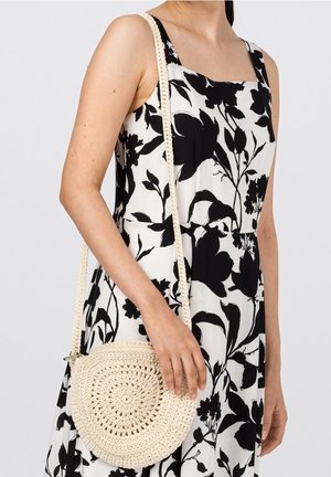 Vrouw in een zwart-witte bloemenjurk die een ronde, roomkleurige gehaakte schoudertas met een lange band vasthoudt.