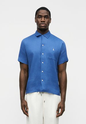 Άνδρας που φοράει μπλε πουκάμισο με κοντά μανίκια και κουμπιά, με ένα μικρό λευκό λογότυπο στην τσέπη του στήθους και λευκό παντελόνι με κορδόνι, στέκεται κοιτώντας μπροστά.