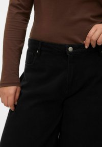 Zwarte denim broek met een hoge taille, voorzien van voorzakken en een metalen knoopsluiting, gecombineerd met een nauwsluitend bruin shirt met lange mouwen.