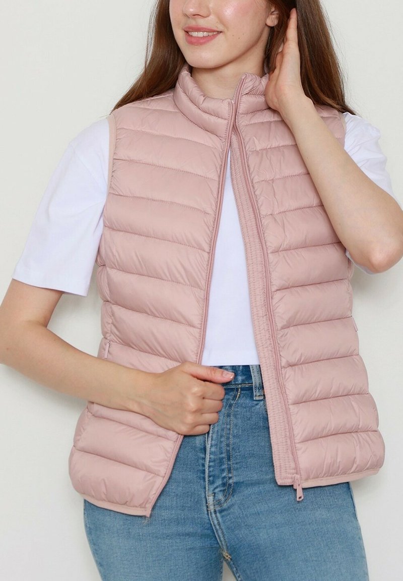 Gilet rembourré rose clair avec un col montant, fermeture zippée et design matelassé vertical, porté sur un t-shirt blanc et un jean bleu.