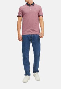 Rode polo shirt met een marineblauwe kraag en gestreepte accenten, gecombineerd met blauwe jeans en witte sneakers. De stof is textuurrijk en heeft een casual pasvorm.