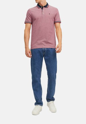 Rode polo shirt met een marineblauwe kraag en gestreepte accenten, gecombineerd met blauwe jeans en witte sneakers. De stof is textuurrijk en heeft een casual pasvorm.