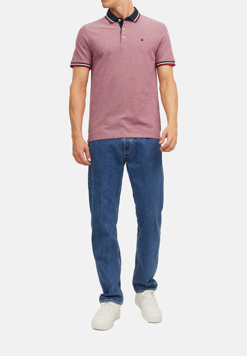 Rode polo shirt met een marineblauwe kraag en gestreepte accenten, gecombineerd met blauwe jeans en witte sneakers. De stof is textuurrijk en heeft een casual pasvorm.