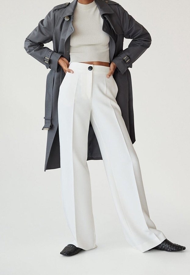 Personne portant un crop top blanc, un pantalon blanc taille haute à jambes larges, des ballerines noires et un trench-coat gris foncé, les mains sur les hanches.