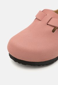 Zoccolo rosa in tessuto con punta arrotondata, dotato di lati traforati e una suola nera a contrasto. Il materiale sembra morbido con una finitura opaca.