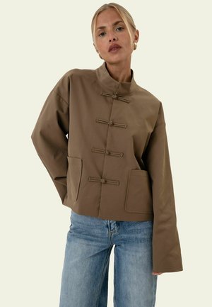 Kvinde iført en brun jakke med frølukninger og frontlommer kombineret med lyseblå jeans mod en ensfarvet baggrund.