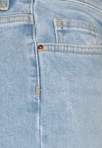 Tecido de denim azul claro com costura visível em amarelo, um pequeno botão de cobre e leve desgaste próximo à costura.