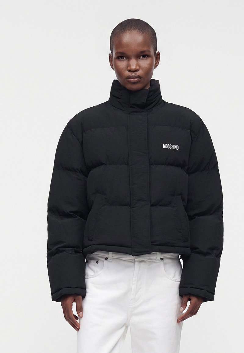Personne portant une doudoune noire Moschino avec col montant et pantalon blanc, debout devant un fond clair uni.