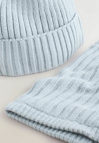 Světle modrý pletený kulich s žebrovaným vzorem a ohrnutým okrajem, doplněný o odpovídající žebrovaný šátek ve stejné barvě a textuře.