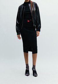 Femme portant une veste bomber en cuir noir, une jupe midi noire à boutons avec une étiquette rouge, un haut noir, des chaussettes en imprimé serpent et des talons ouverts noirs.