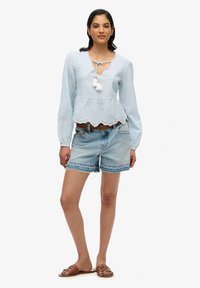 Top peplum a rayas en azul claro, con dobladillo ondulado y cuello con lazo; combinado con pantalones cortos de mezclilla deshilachados y sandalias marrones.