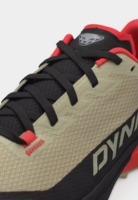 Athletic shoe with beige mesh upper, black laces, red accents, and a logo on the tongue and side reading "DYNA".