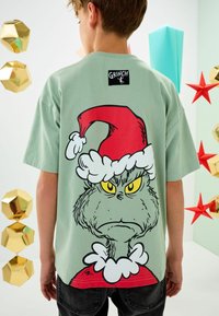 Zaļa īsām piedurknēm T-krekls ar lielu karikatūras Grinču, kas valkā sarkano Ziemassvētku ķiveri un balto kažokādu apkaklē. Melns "GRINCH" uzraksts aizmugurē.