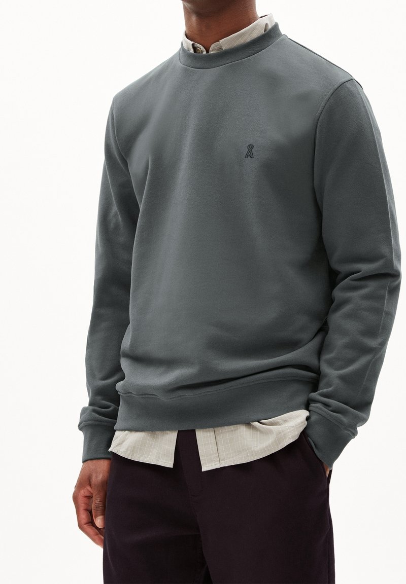 Homme portant un sweat-shirt gris foncé sur une chemise beige à boutons, les mains dans les poches d’un pantalon foncé, sur fond blanc.