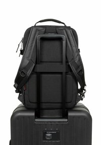 Černý látkový batoh s síťovaným zadním panelem, polstrovanými popruhy a popruhem na zavazadla, umístěný na pevném kufru. Viditelný kovový herní prvek.