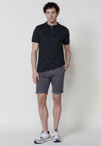 Crna polo majica s geometrijskim uzorkom, u kombinaciji s sivim hlačama do koljena, upotpunjena bijelim sportskim cipelama. Vidna je kovrčava kosa.
